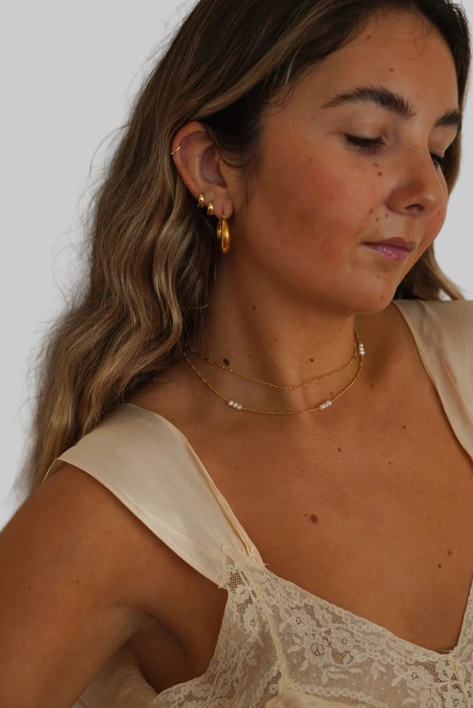 Woman wearing gold earrings and necklaces against a neutral background