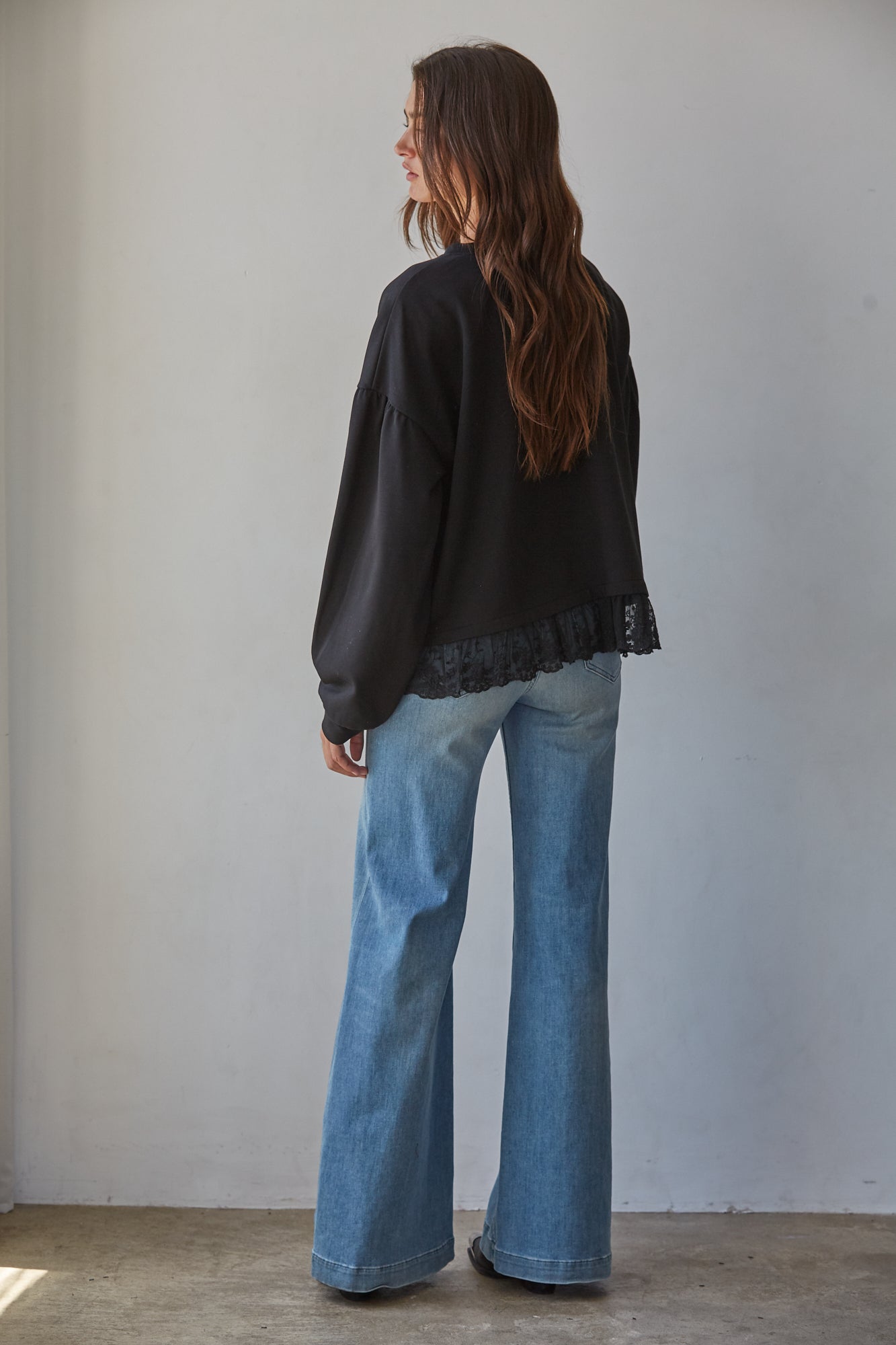 A woman modeling a black long-sleeve crewneck cotton sweatshirt with a lace hem, paired with wide-leg blue jeans.