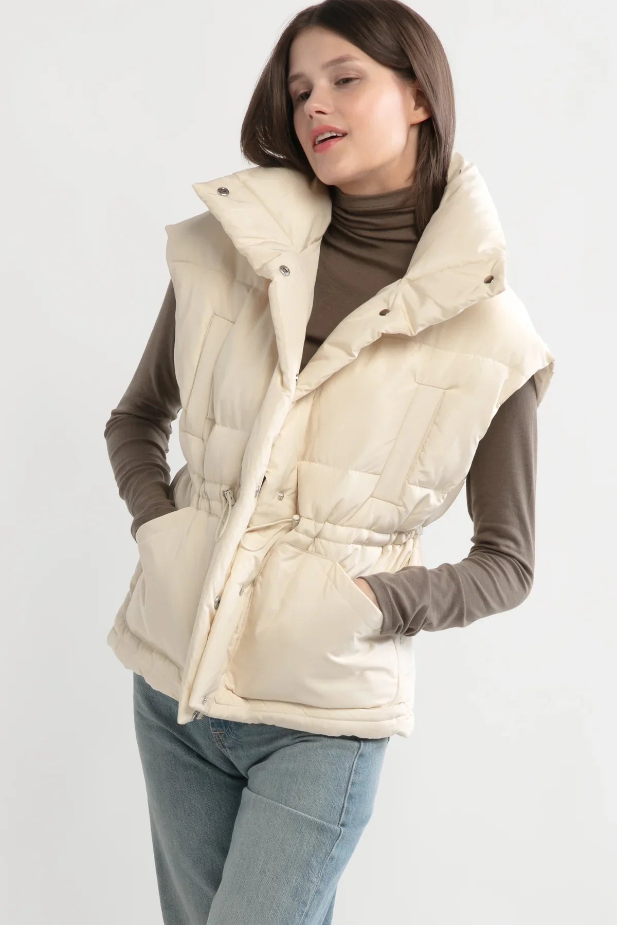 Model wearing a cream puffer vest with high collar snap closure with toggle pull and turtleneck.