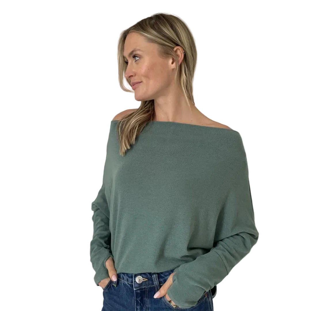 A woman wearing a muted sage green off-the-shoulder knit sweater and blue jeans, shown from the waist up against a plain white background.
