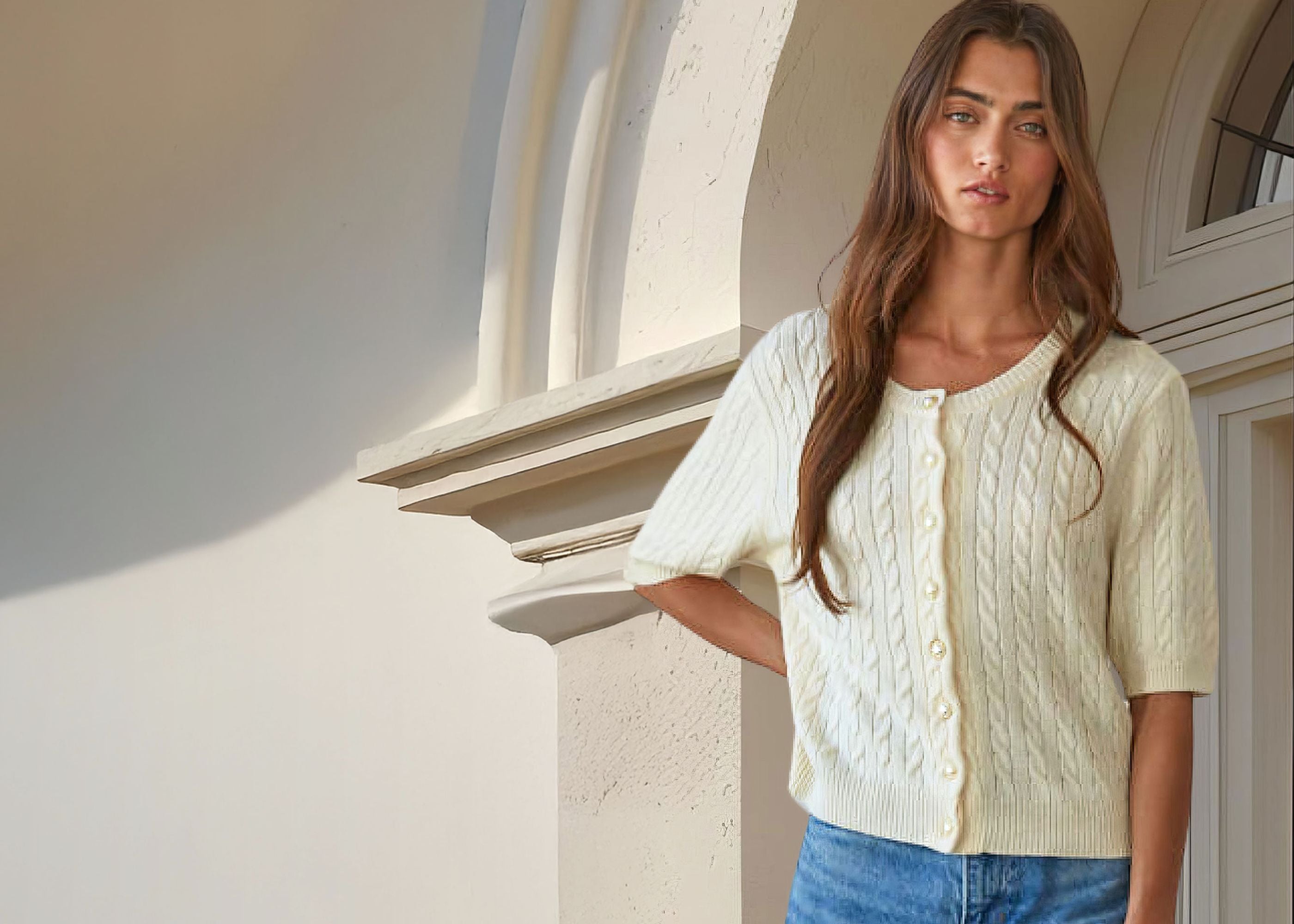 Woman wearing a light-colored cable knit cardigan and blue jeans standing against a neutral wall.