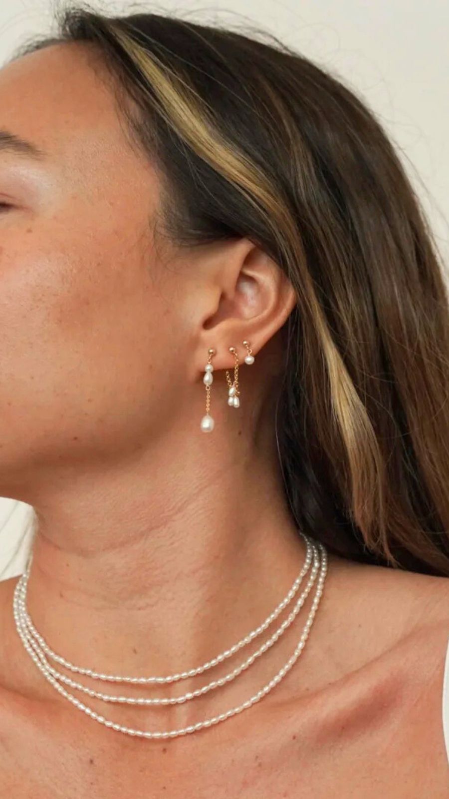 Close-up of a woman wearing pearl earrings and a necklace against a neutral background